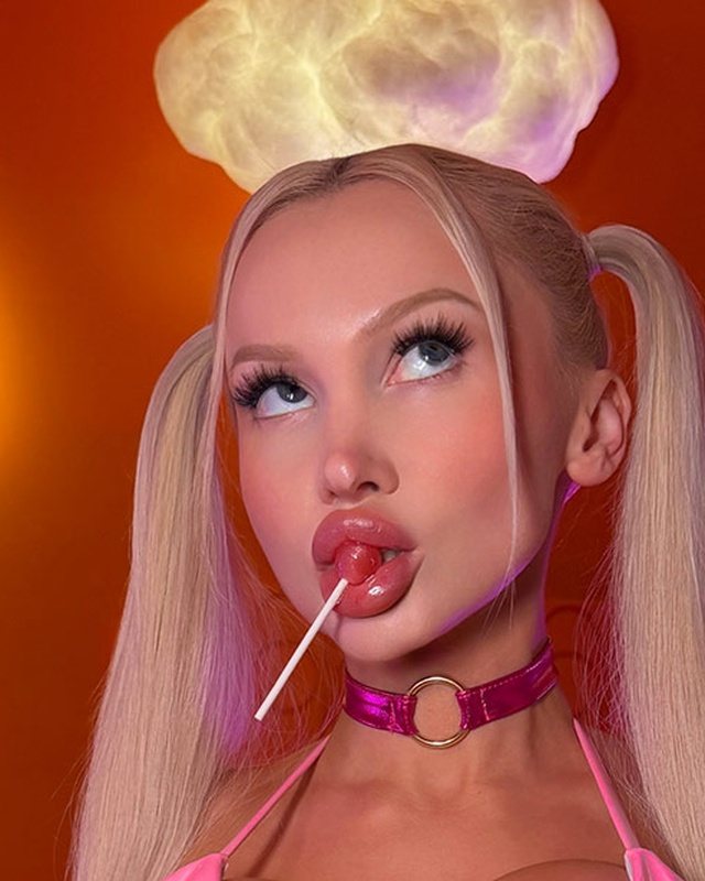 Bimbo look with platinum pigtails, a pink collar and a lollipop against an orange backdrop.